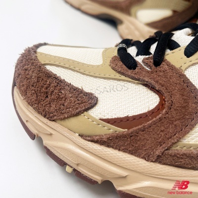 Ténis New Balance bege com detalhes castanhos e atacadores pretos, fundo branco.