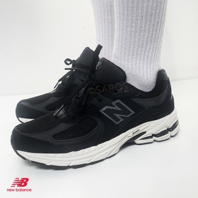 Tênis New Balance preto com sola branca e meias brancas em fundo branco.