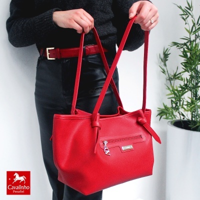 Mala roja de senhora com alças e fecho dianteiro, usada por pessoa com roupa preta junto a planta.