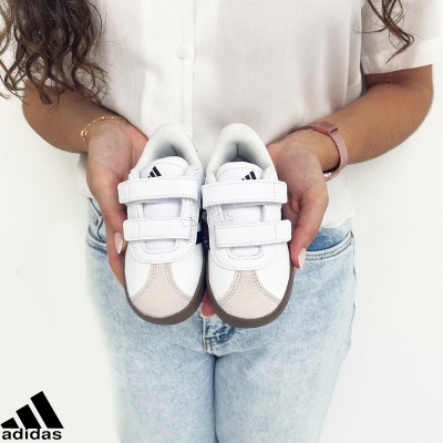 Sapatilhas infantis Adidas brancas com fecho de velcro seguradas por pessoa com camisa branca e calças de ganga