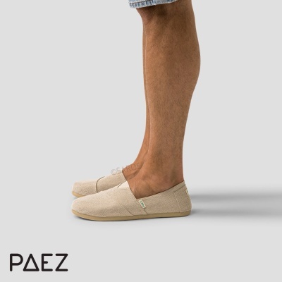 Alpercatas Paez bege com sola plana em pernas de homem com calças curtas no fundo cinzento.