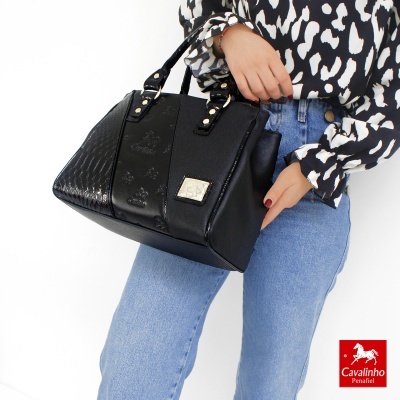 Mala preta com textura croco e detalhes metálicos, segurada por pessoa com camisa preta e branca e jeans azuis