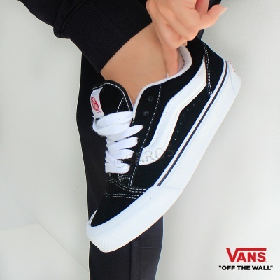 Ténis Vans pretos com padrão branco e sola branca, usados por pessoa com calças pretas