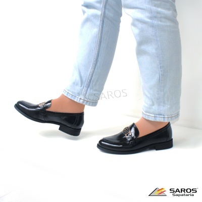 Sapatos mocassins pretos em verniz com fivela dourada e calças de ganga claras.