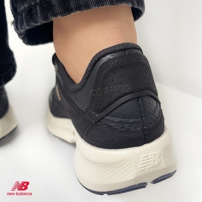 Ténis New Balance pretos com sola branca, visto de trás num pé com calças pretas.