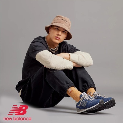 Jovem sentado com roupa preta, ténis New Balance azuis e chapéu bege