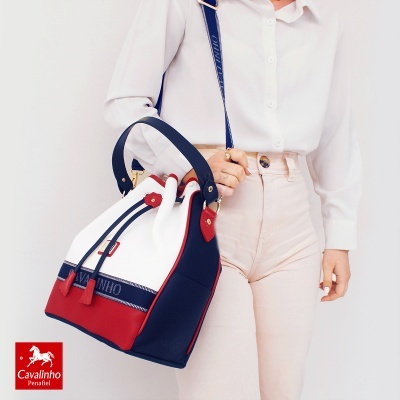 Mala branca, vermelha e azul escuro em saco com alças e tiracolo, com modelo a usar camisa branca e calças bege.