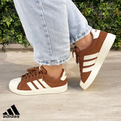 Ténis Adidas castanhos com riscas brancas usados com calças de ganga claras em chão de madeira