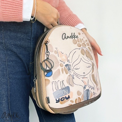 Mochila pequena bege e rosa com estampado de menina e texto, chaveiro metálico, segurada por pessoa com camisola rosa e jeans.