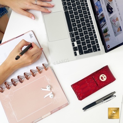 Agenda rosa com argolas metálicas, caneta, auscultadores e estojo vermelho em mesa branca ao lado de portátil.