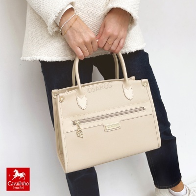 Mala de mão bege em pele sintética com fecho de correr e chaveiro metálico segurada por pessoa com casaco branco e calças pretas