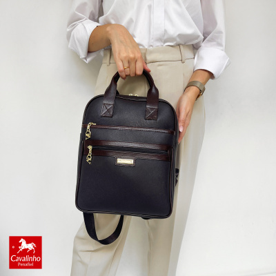 Mochila preta e castanha em pele sintética com dois bolsos e alças, segurada por pessoa com camisa branca e calças claras.