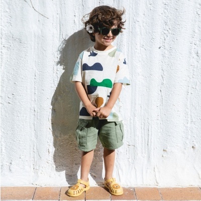 Menino com t-shirt estampada, calções cáqui e sandálias amarelas junto a parede branca.