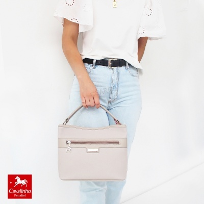 Mala bege claro com alça, fecho zipper frontal, segurada por mulher com calças azul claro e t-shirt branca