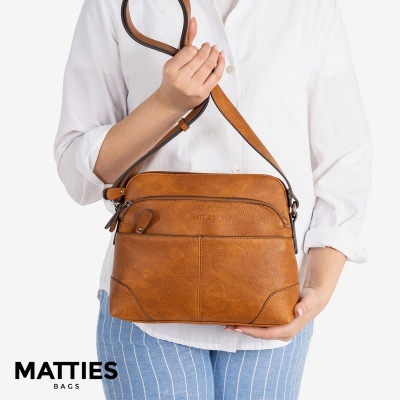 Mala castanha em couro tipo saco com alça ajustável, alguém segura a mala vestindo camisa branca e calças riscas azuis.