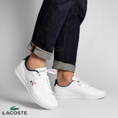 Sapatilhas brancas Lacoste com detalhes azul-marinho e logo, calças de ganga dobradas.