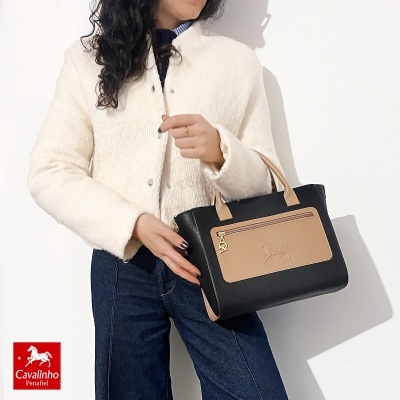 Mala de mão Cavalinho Penafiel preta e bege com fecho dourado segurada por modelo com casaco branco e jeans.