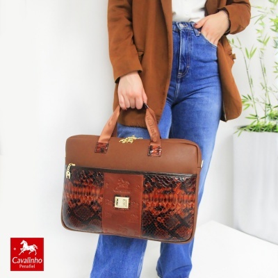 Mala castanha em pele com padrão tipo pele de cobra segurada por pessoa com casaco castanho e calças azuis