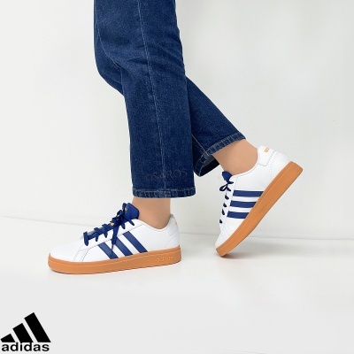 Ténis Adidas brancos com riscas azuis e sola castanha usados com calças de ganga escuras