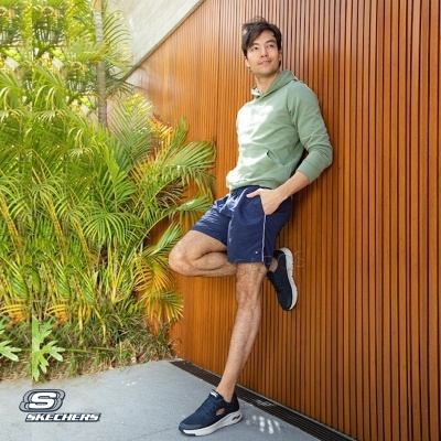 Homem com outfit casual encostado em parede de madeira junto a plantas.