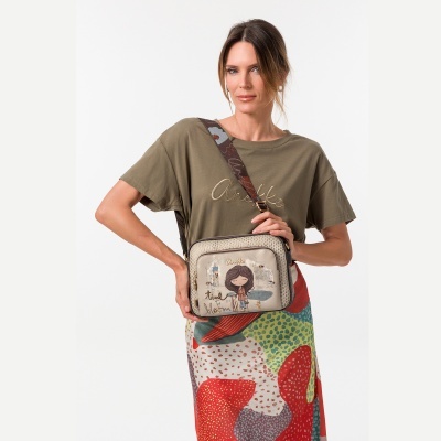 Mulher com t-shirt kaki e saia colorida segurando mala bege com design de personagem e texto Anekke