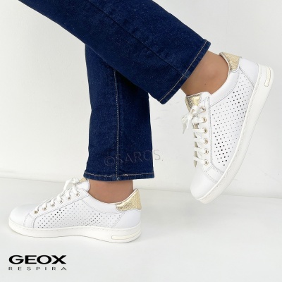 Ténis brancos com detalhes dourados e perfurações, usados com calças azuis.