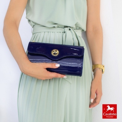 Carteira azul escuro em verniz com fecho dourado, segurada por mulher em vestido verde claro plissado