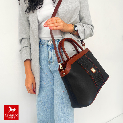 Mala preta e castanha com padrão central e alças, usada por mulher com blazer cinza e jeans azuis.