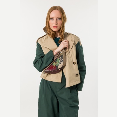Mulher com macacão verde e casaco bege usa bolsa tipo pochete bege com padrão floral e texto Anekke.