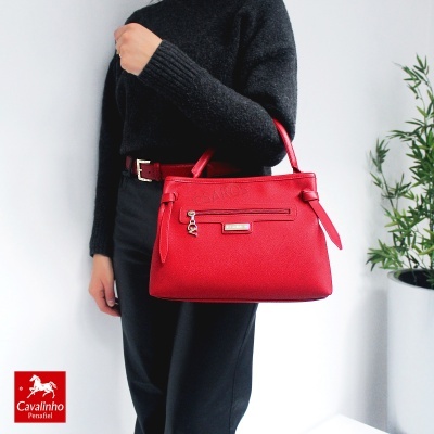 Mala de mão vermelha com fecho e placa cavalinho, mulher de sweater preto e calças escuras junto a planta em vaso branco