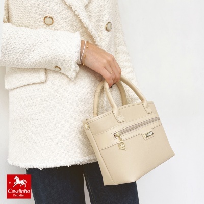 Mala bege de mão com fecho e pendente dourados, casaco bege texturado e botões, mão segurando a mala