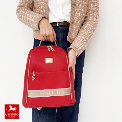 Mochila vermelha Cavalinho com detalhes bege e fechos dourados. Pessoa com casaco castanho claro e calças azuis escuras segura a mochila.