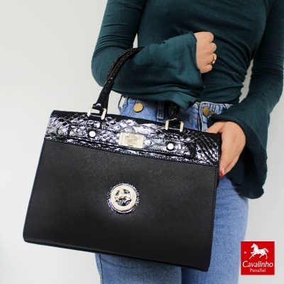 Mala preta com textura croco e detalhes metálicos, segurada por pessoa com camisola verde e jeans