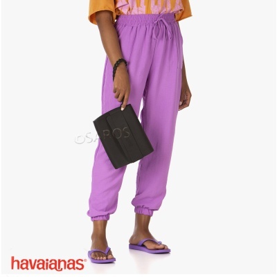 Pessoa com calças lilás, chinelos lilás Havaianas, camiseta colorida e bolsa preta