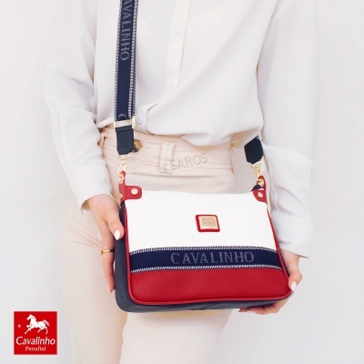 Mala Cavalinho branca, vermelha e preta com alça azul com texto, usada por pessoa com camisa branca e calças bege