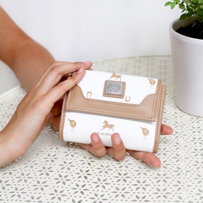 Carteira branca e castanho claro com padrões dourados e placa metálica, segurada na mão sobre mesa branca perfurada, com vaso branco e planta ao lado.