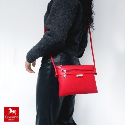 Mala de ombro vermelha em tecido com fecho e pendente metálico, alça ajustável, usada por pessoa vestindo roupa escura com fundo branco