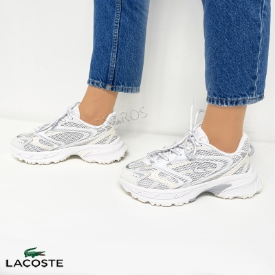 Ténis brancos Lacoste com detalhes cinza e bege, usados com jeans azuis.