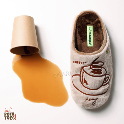 Pantufa felpuda bege com desenho de chávena de café e texto, ao lado de copo de papel virado com café derramado