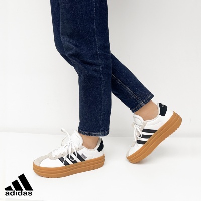 Ténis Adidas brancos com riscas pretas e sola castanha em fundo branco
