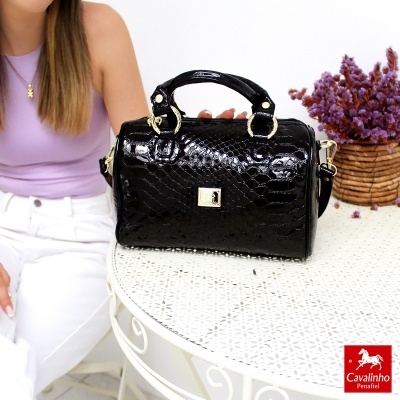 Mala preta de verniz com textura de crocodilo sobre mesa branca com vaso de flores roxas