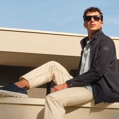 Homem com casaco preto, calças claras e óculos de sol sentado em estrutura exterior.