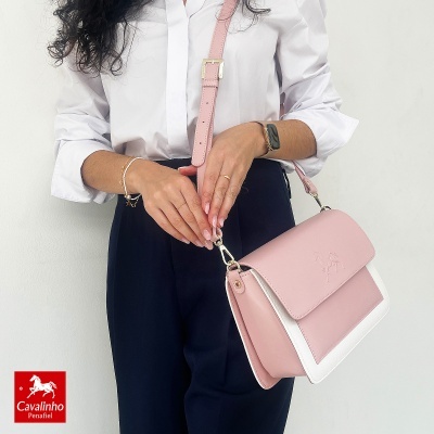 Mala rosa com detalhes brancos, alça rosa, fechos dourados, usada por mulher com camisa branca e calças pretas