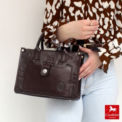 Mala de couro castanho com padrão embossado segurada por pessoa com camisa castanha e calças de ganga.