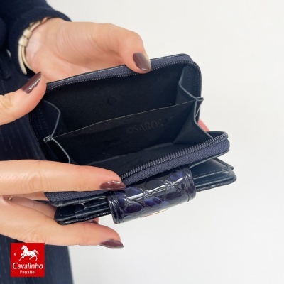 Carteira azul escuro croco com fecho de correr e interior preto com texto SAROS, segurada por mãos femininas.