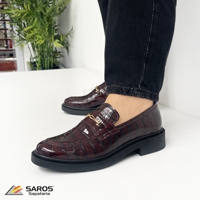 Sapatos tipo mocassim castanho escuro em couro com padrão crocodilo e detalhe metálico dourado, usados por uma pessoa com calças pretas