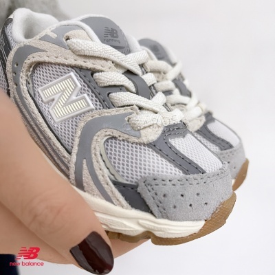 Tênis New Balance cinza, bege e branco para criança em fundo branco, segurados por uma mão com unhas castanho escuro