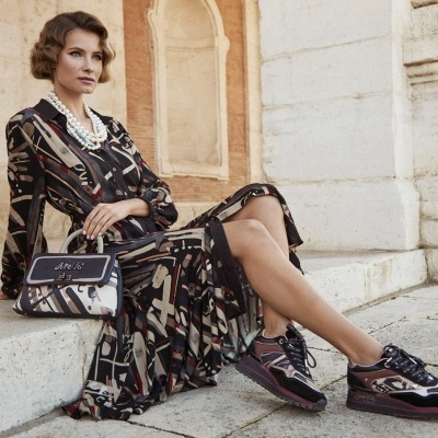 Mulher sentada com vestido estampado e sapatilhas coloridas, mala pequena, degrau de pedra