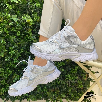 Ténis Nike brancos com detalhes prateados e sola branca, usados por pessoa com calças bege, contra fundo de folhas verdes.