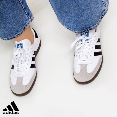 Ténis Adidas brancos com riscas pretas e sola castanha, usados com calças de ganga azuis